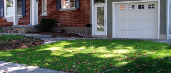 A suburban house with a brick facade, white garage door, and a manicured lawn sprinkled with fallen leaves under a bright, sunny sky.