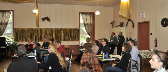 This image shows a group of people seated in rows, attending a meeting or conference in a room with traditional decor and a piano in the corner.