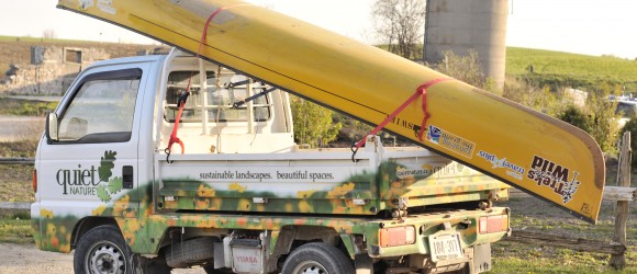 A commercial truck carrying a long yellow canoe is parked outdoors. The truck's livery suggests it belongs to a landscaping company.