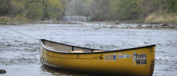 A yellow canoe is tethered in a calm river, surrounded by natural vegetation, with trees and possibly a small waterfall in the background.