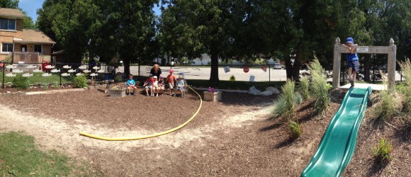 This is a panoramic view of a sunny playground featuring a green slide, people sitting on benches, a sandbox, and a person standing on playground equipment.