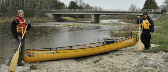 Two people stand by a yellow canoe near a riverbank with paddles in hand, wearing life jackets, and a bridge visible in the background.