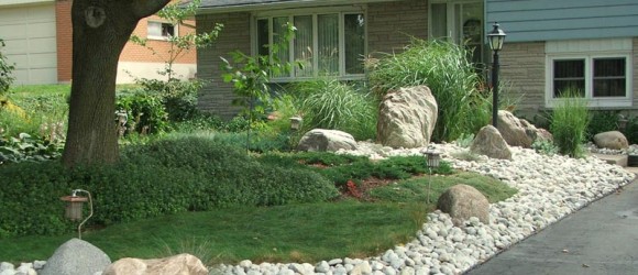 A well-manicured suburban garden with a variety of plants, large decorative rocks, and a pebble path next to a driveway leading to a house.