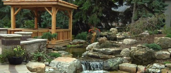 The image shows a serene garden with a wooden gazebo, a rock-lined pond, cascading waterfalls, lush plants, and a few blooming flowers.