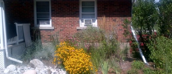 The photo shows a brick house with a white porch rail, window air conditioning units, a well-kept garden with yellow flowers, and a sunny sky.