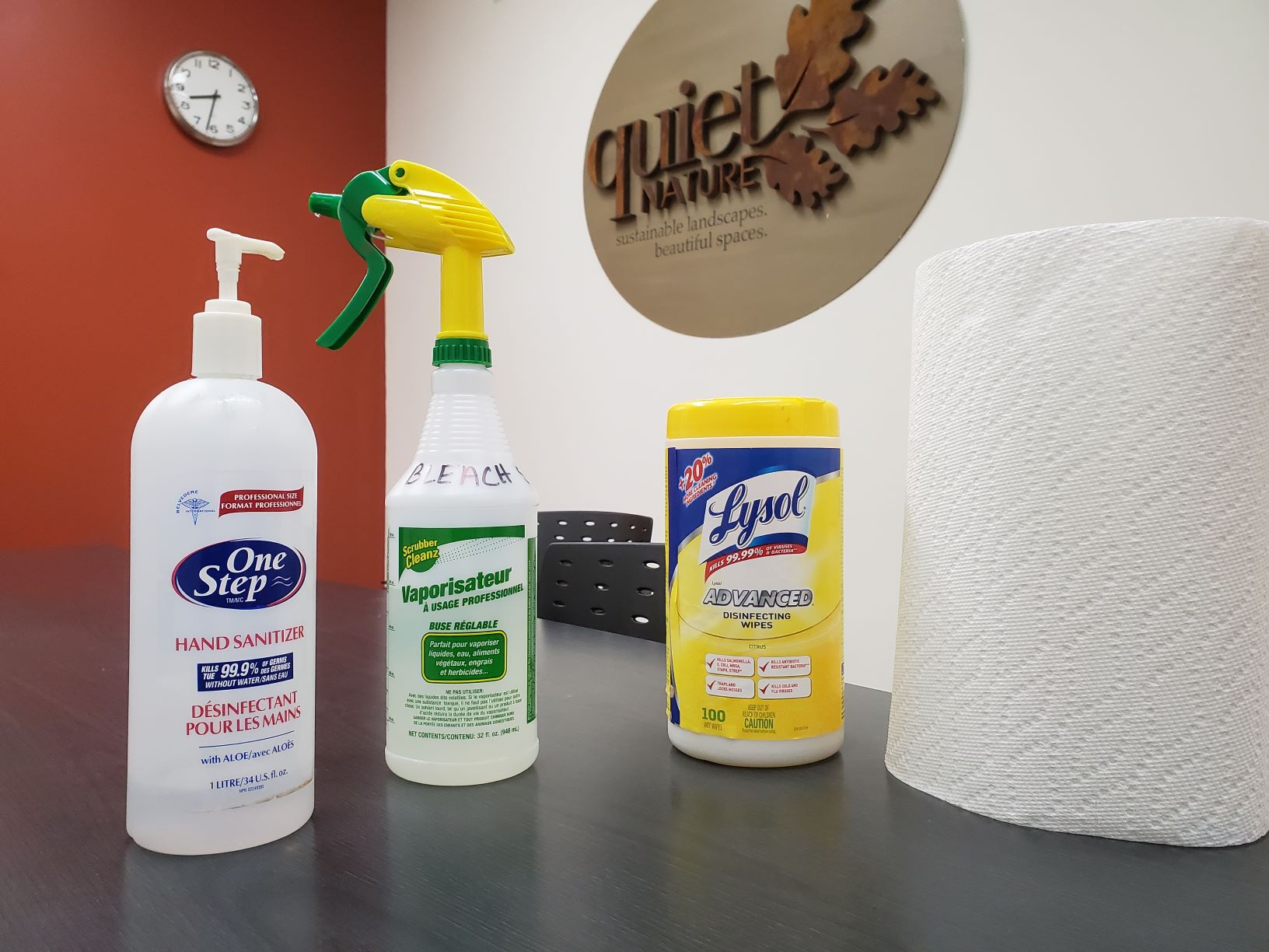 This image depicts a collection of cleaning supplies on an office table, including hand sanitizer, a spray bottle of bleach, disinfecting wipes, and a paper towel roll.