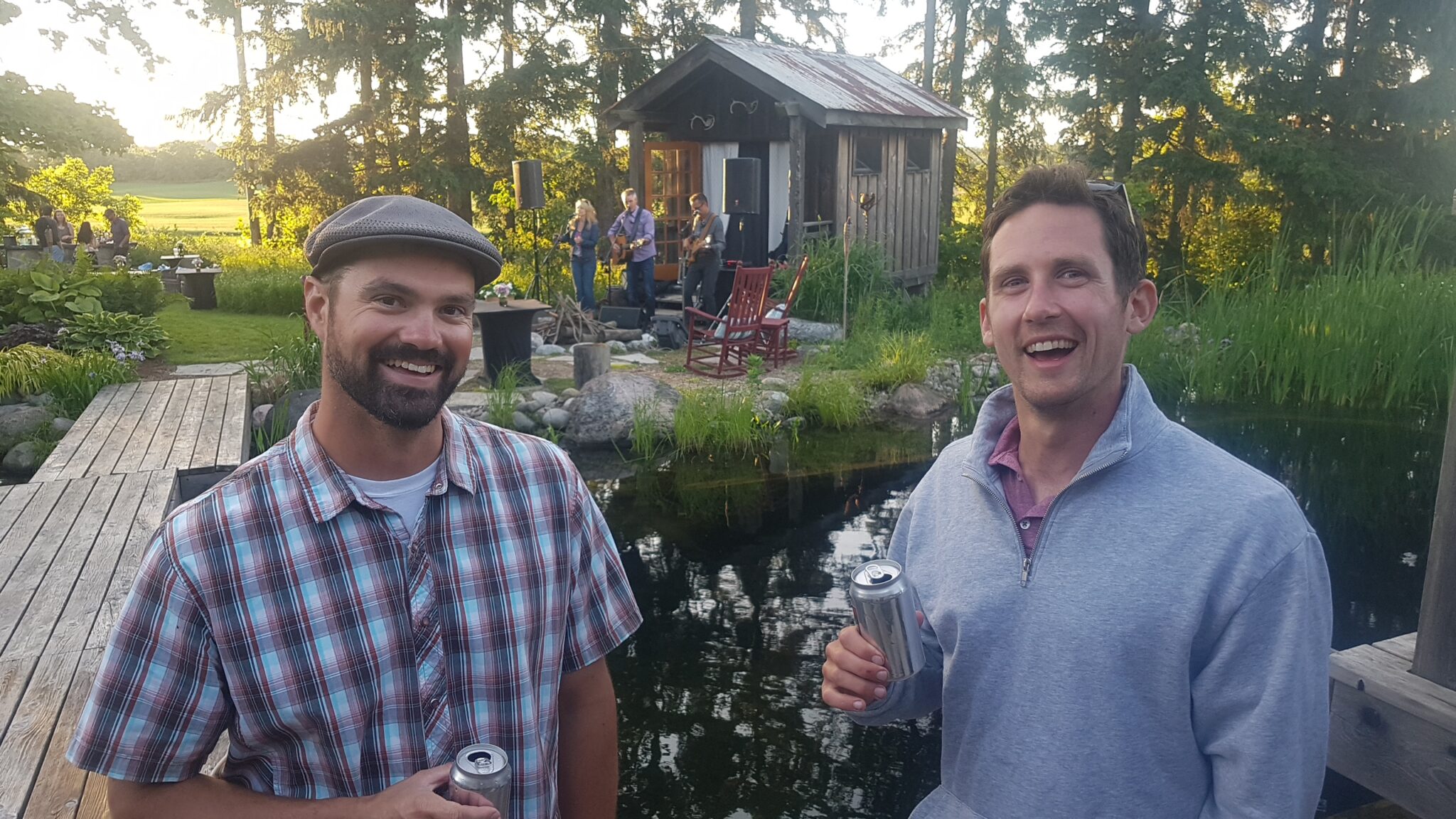 Two people are smiling at the camera, holding beverages, with a rustic shed and a small band performing in the background near a pond.