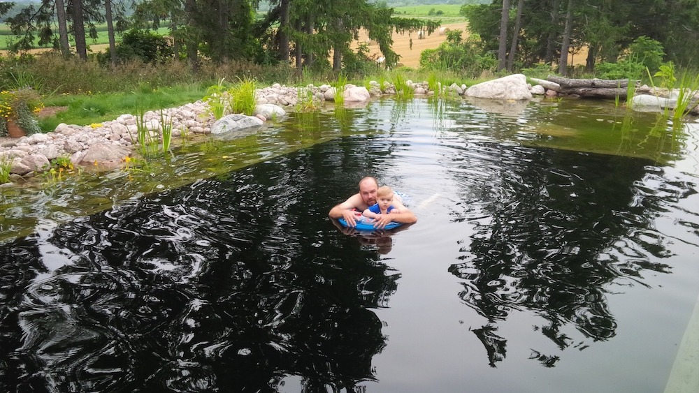 A person and a child are enjoying time together in a natural pond surrounded by rocks and greenery, with fields and trees in the background.