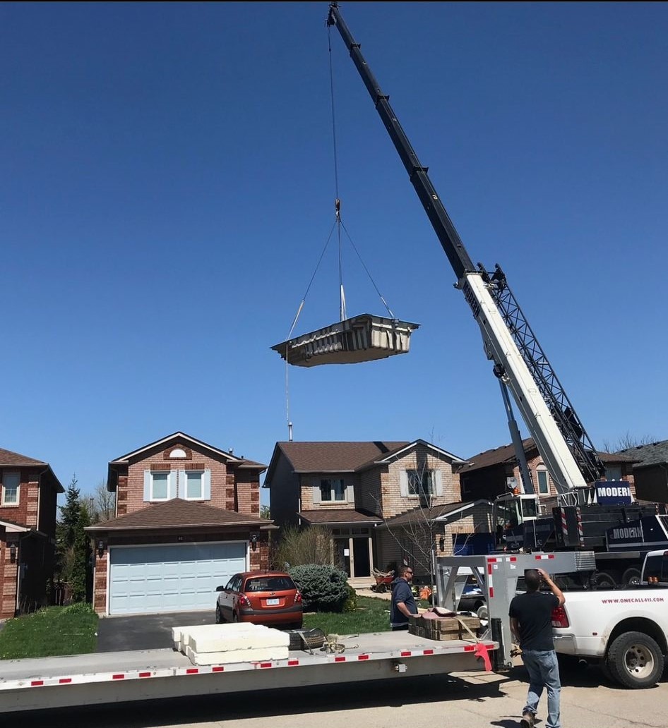 A crane hoists a large concrete slab above a residential street, while several people oversee the operation on a sunny day near suburban homes.