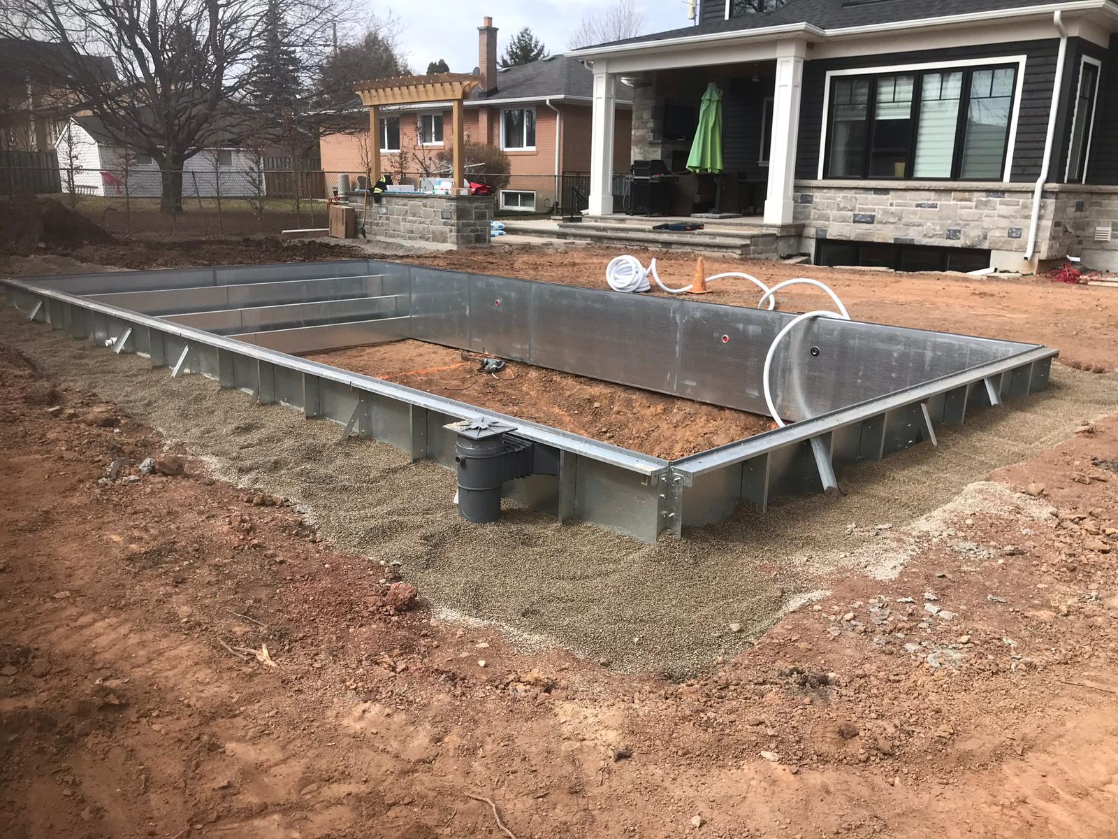 The image shows a galvanized steel frame foundation for a construction project, possibly a deck or small structure, with a residential setting in the background.