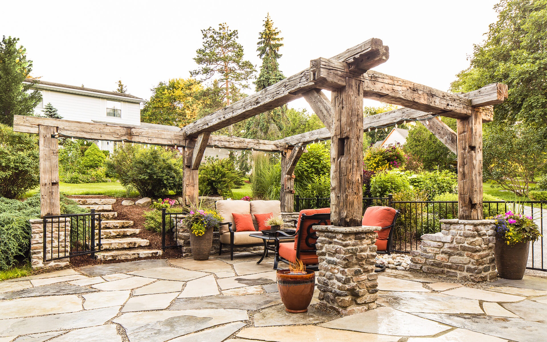 An inviting outdoor patio with a rustic wooden pergola, stone fireplace, comfortable seating, and lush greenery surrounding a flagstone floor.