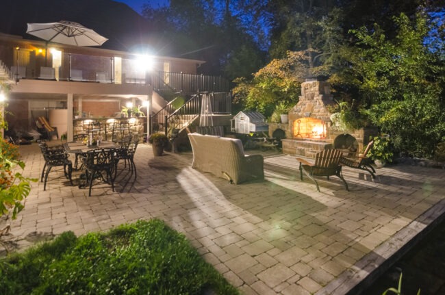 An illuminated backyard at dusk featuring a fireplace, outdoor furniture, a dining set, and a two-story house with a balcony. Trees enclose the space.