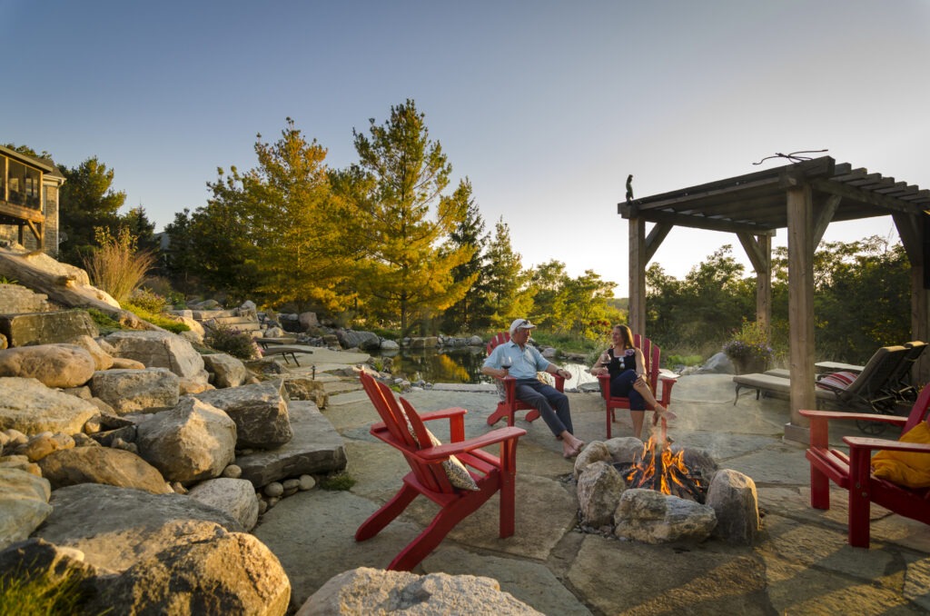 Two people are sitting in red Adirondack chairs by a fire pit, enjoying a tranquil outdoor setting during sunset with trees and a pond nearby.