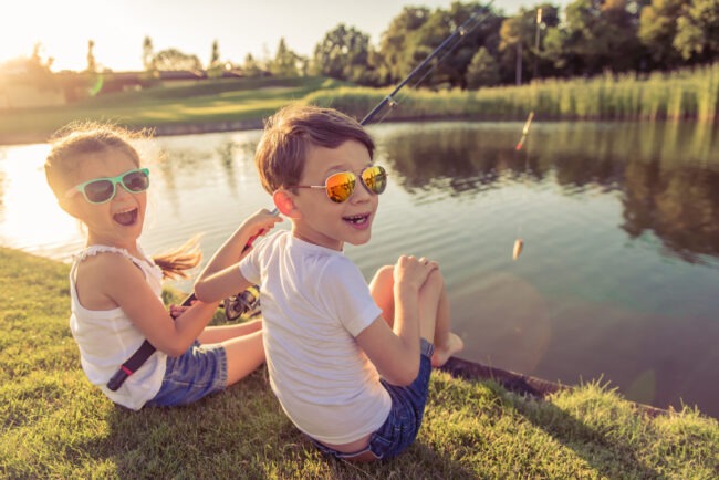 Two children sit by a lakeside at sunset, fishing and laughing together. They wear sunglasses and look relaxed, enjoying the outdoor activity.