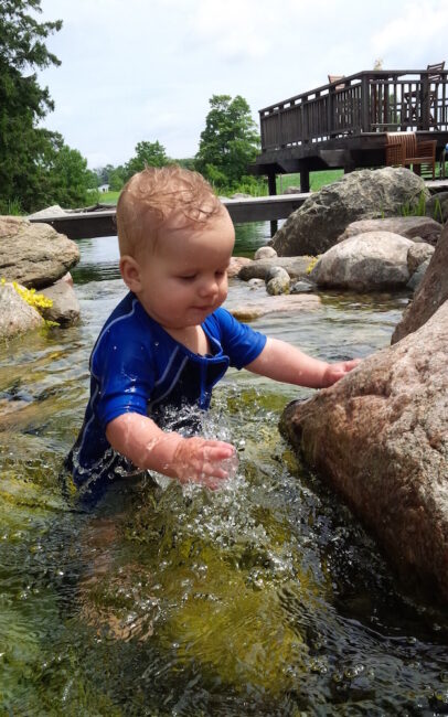 A baby in a blue swimsuit is playing with water between rocks, with a wooden bridge and lush trees in the background on a sunny day.