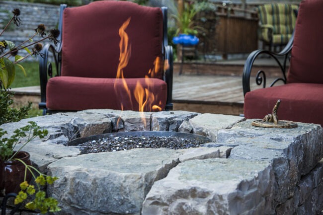 A backyard setting with a stone fire pit ablaze, surrounded by cozy chairs with red cushions, plants, and a wooden deck in the background.