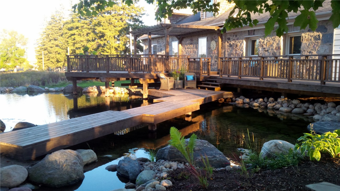 A tranquil setting featuring a wooden deck over a pond with rocks, leading to a stone building with large windows, surrounded by lush greenery at sunset.