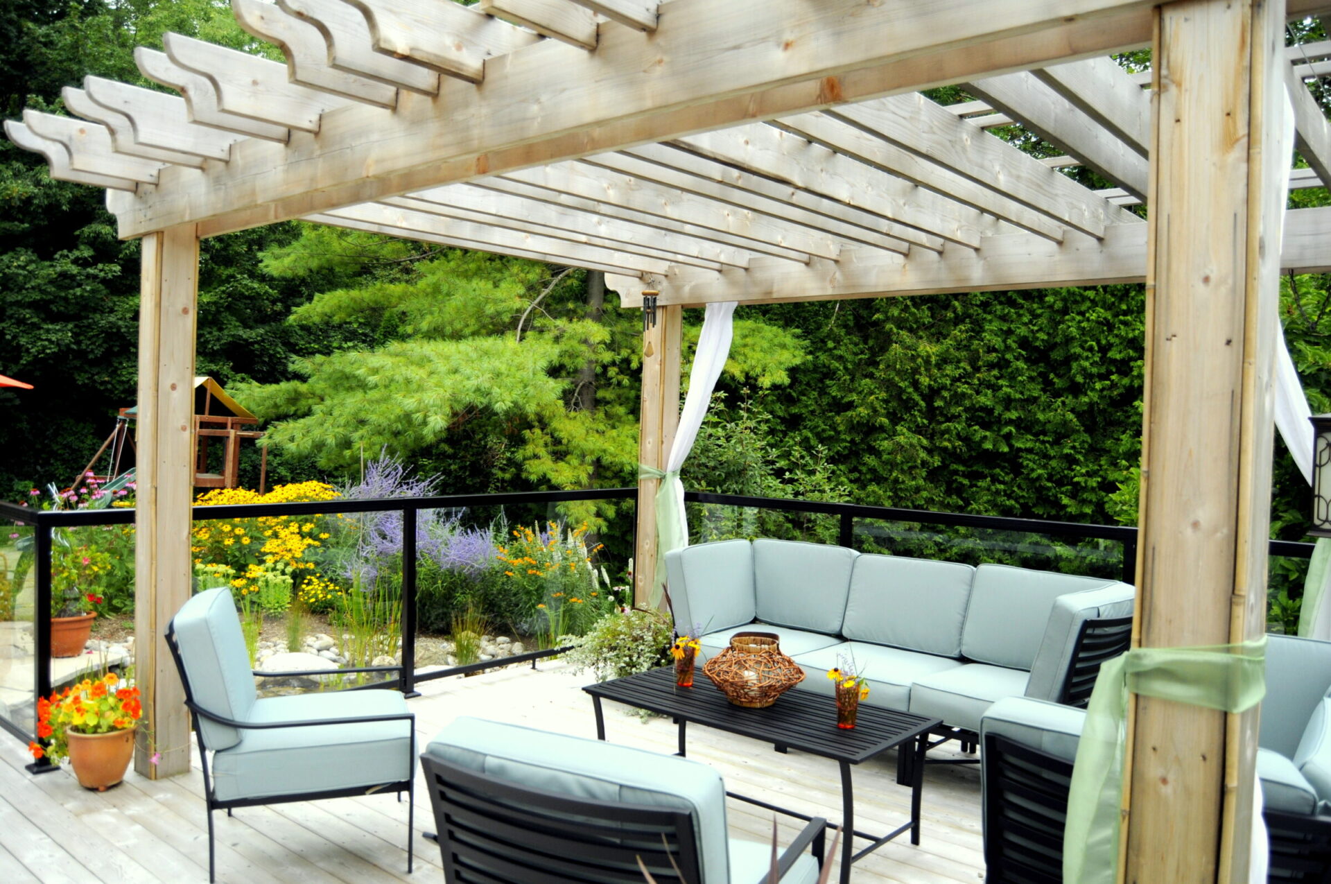 A cozy outdoor patio with a wooden pergola, sectional sofa, and colorful garden in the background, inviting relaxation and outdoor living.