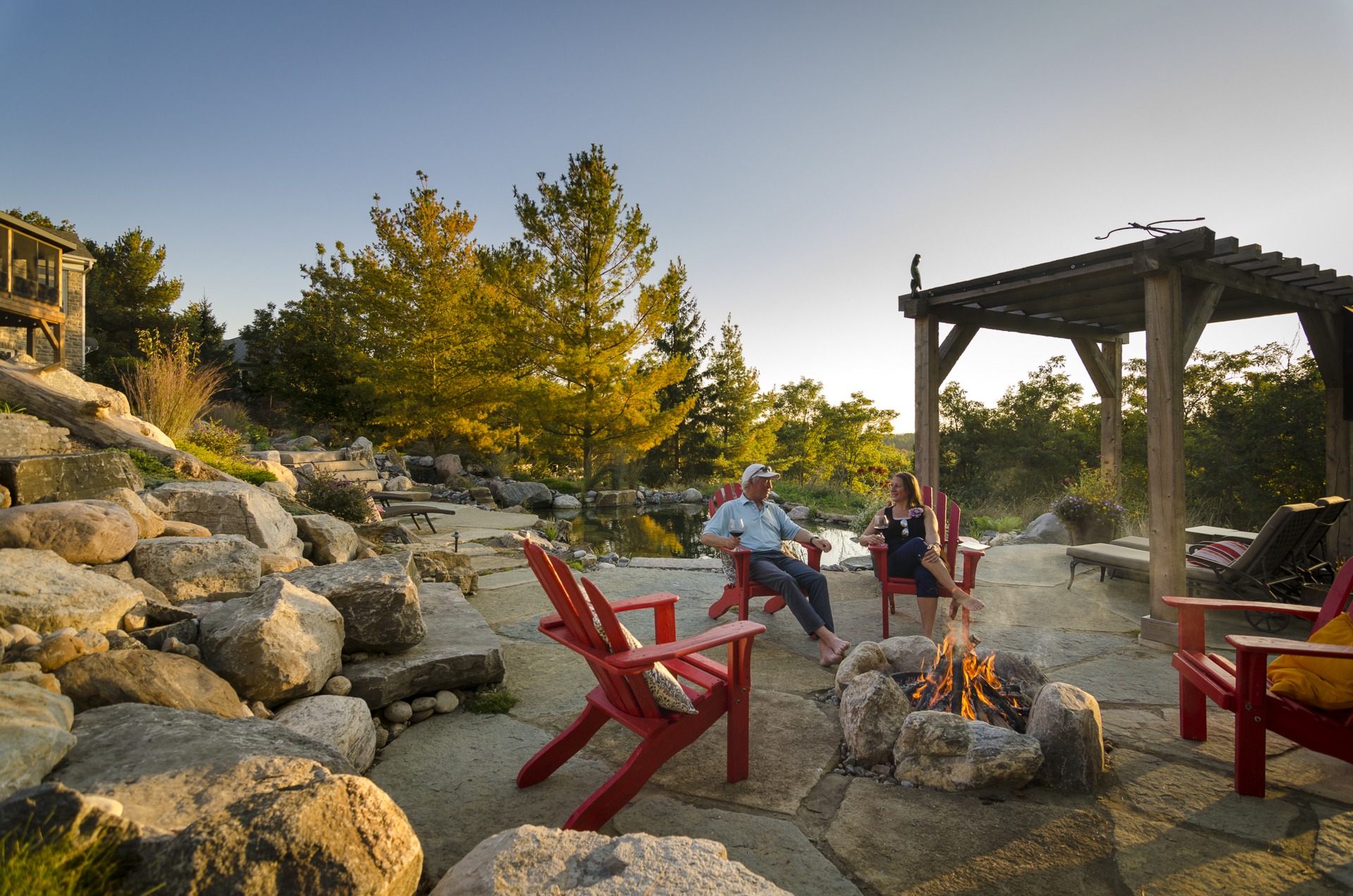 Two people sit on red chairs by a fire pit, enjoying conversation in a serene outdoor setting with trees, rocks, and a wooden pergola.