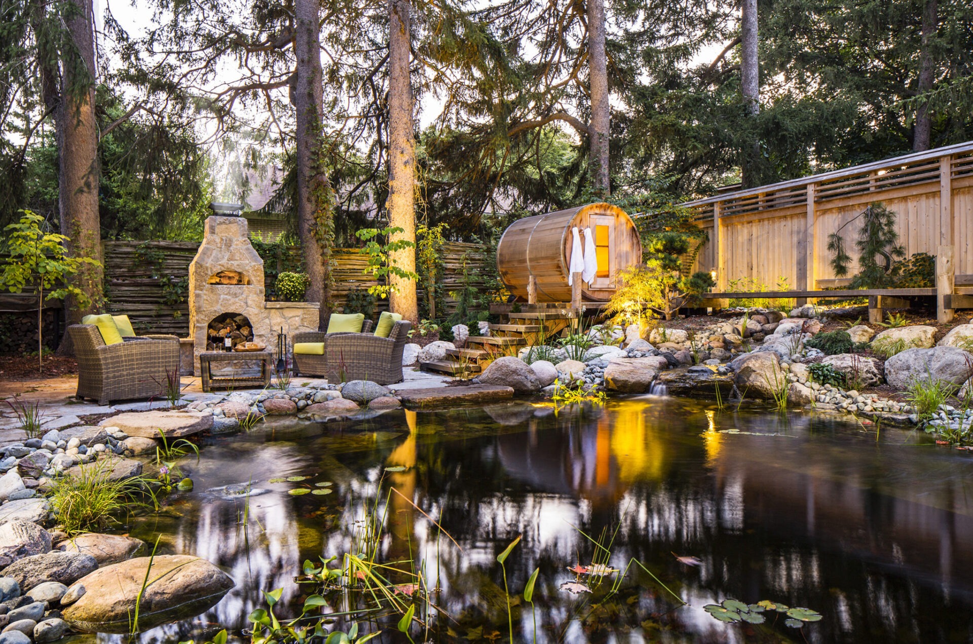 A serene outdoor space with a fireplace, seating area, reflective pond, barrel sauna, lush trees, and ambient lighting for a tranquil atmosphere.