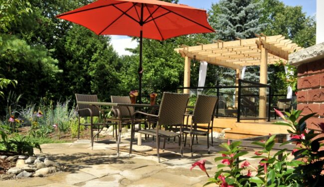 A tranquil garden patio with a large red umbrella, wicker chairs around a table, a pergola, lush greenery, and blooming flowers in bright sunlight.