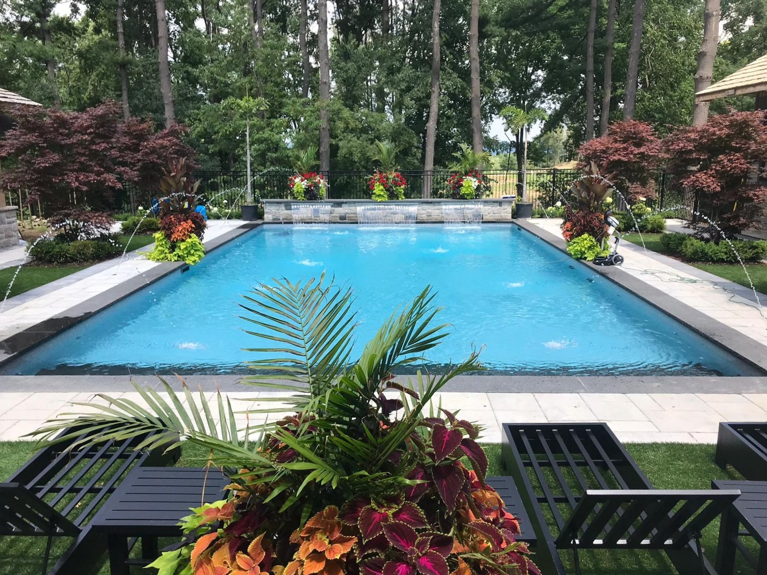 An outdoor swimming pool is surrounded by lush landscaping, colorful flowers, and tall trees, with elegant water features and patio furniture.