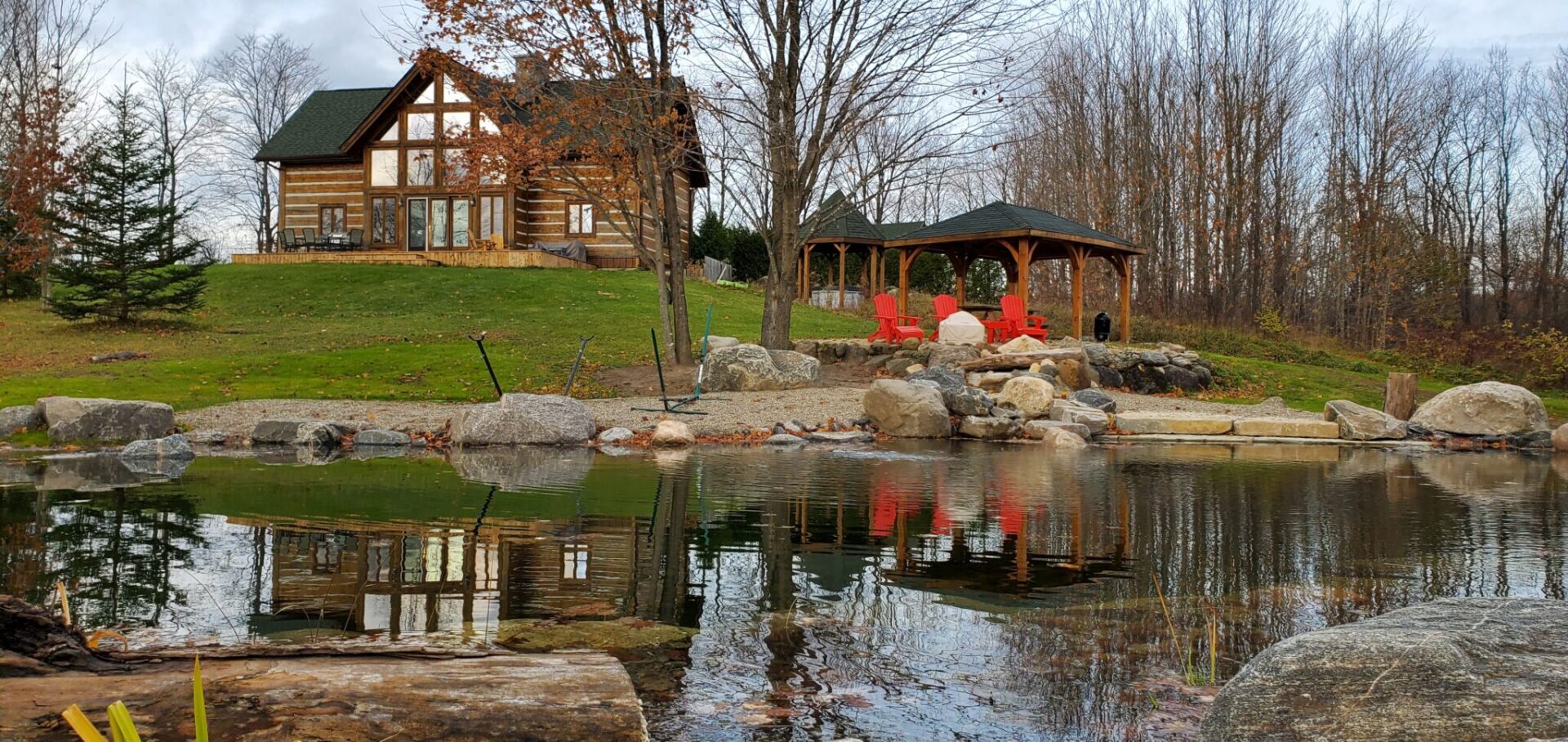 A tranquil setting featuring a log cabin overlooking a pond with a gazebo and red chairs nearby, surrounded by rocks and autumn foliage.