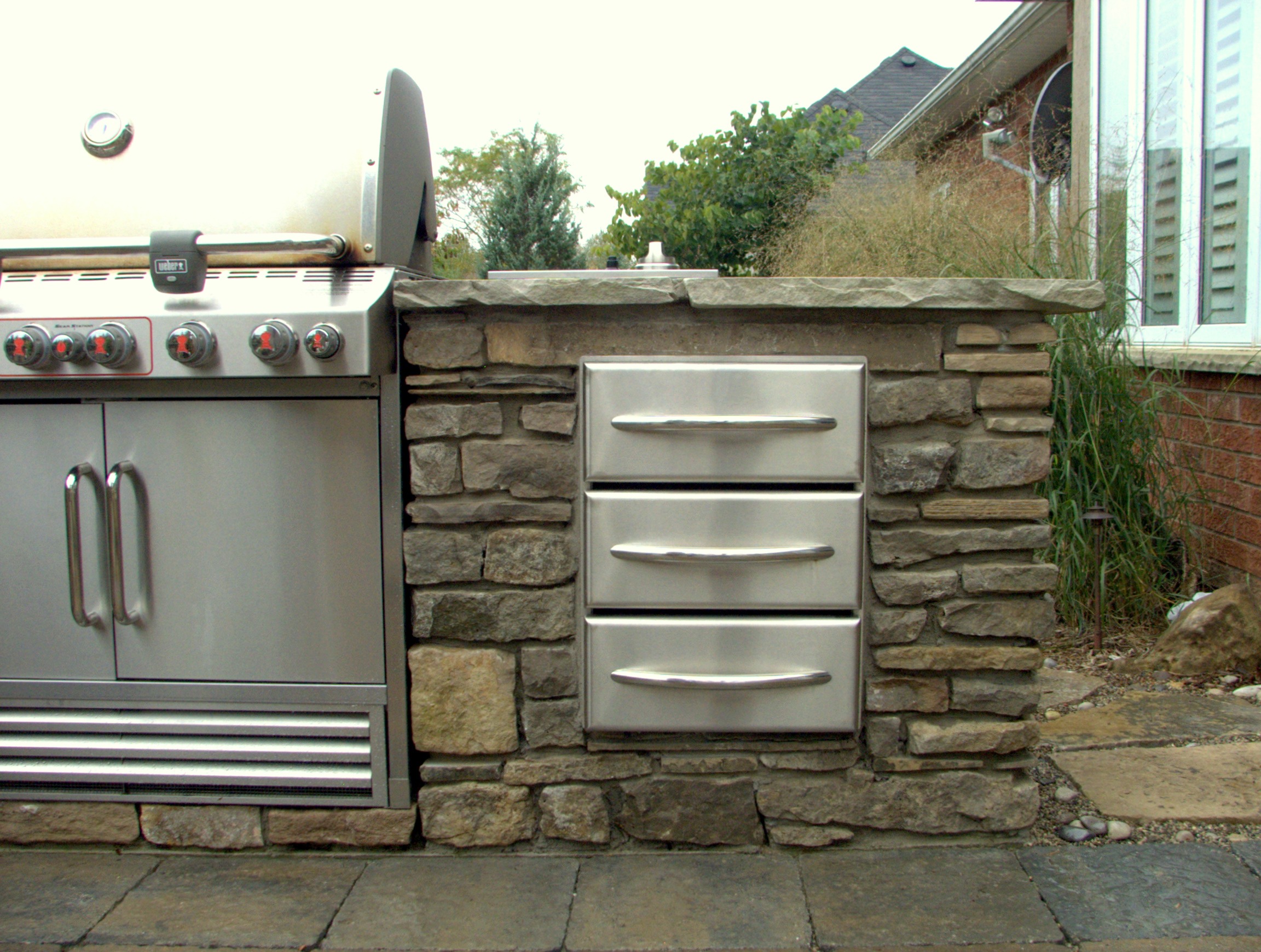 An outdoor kitchen space featuring a stainless steel grill and drawers built into a stone structure, with a house and plants in the background.