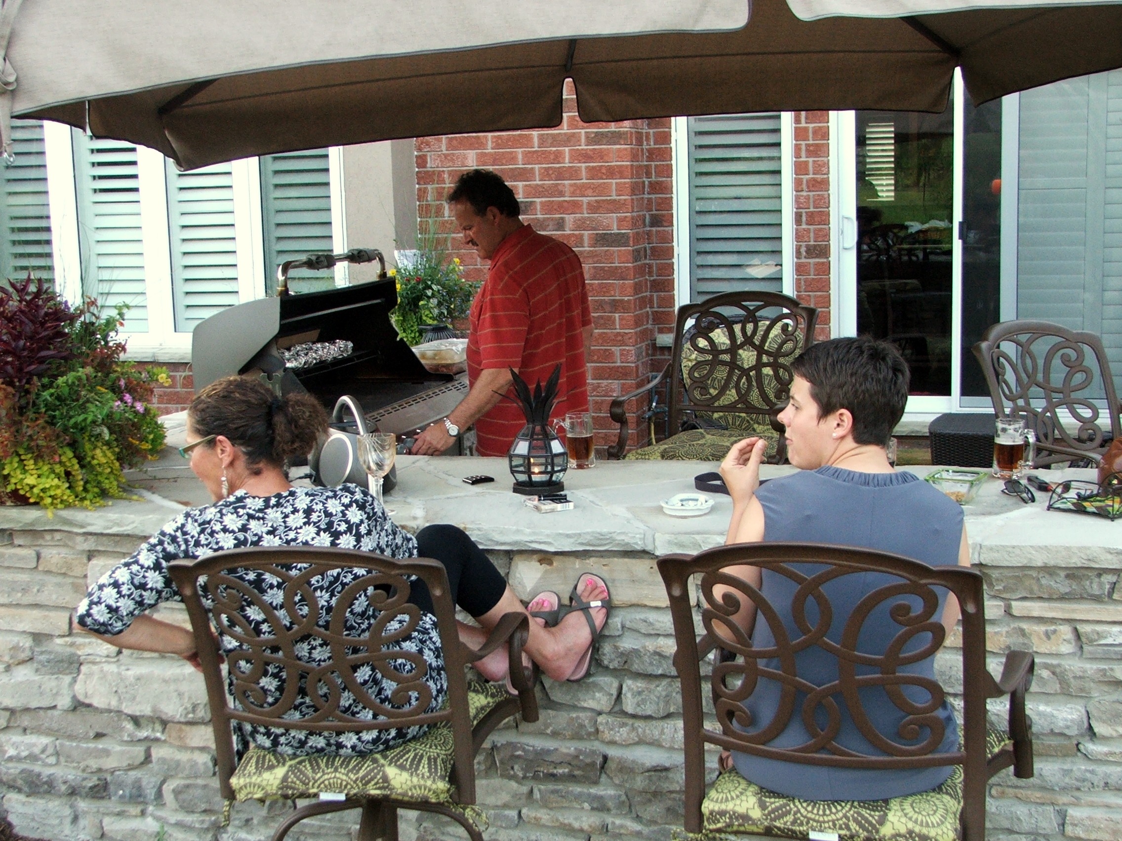 Two people are seated outdoors at a stone bar; a person is grilling in the background. Plants, patio furniture, and a house are visible.