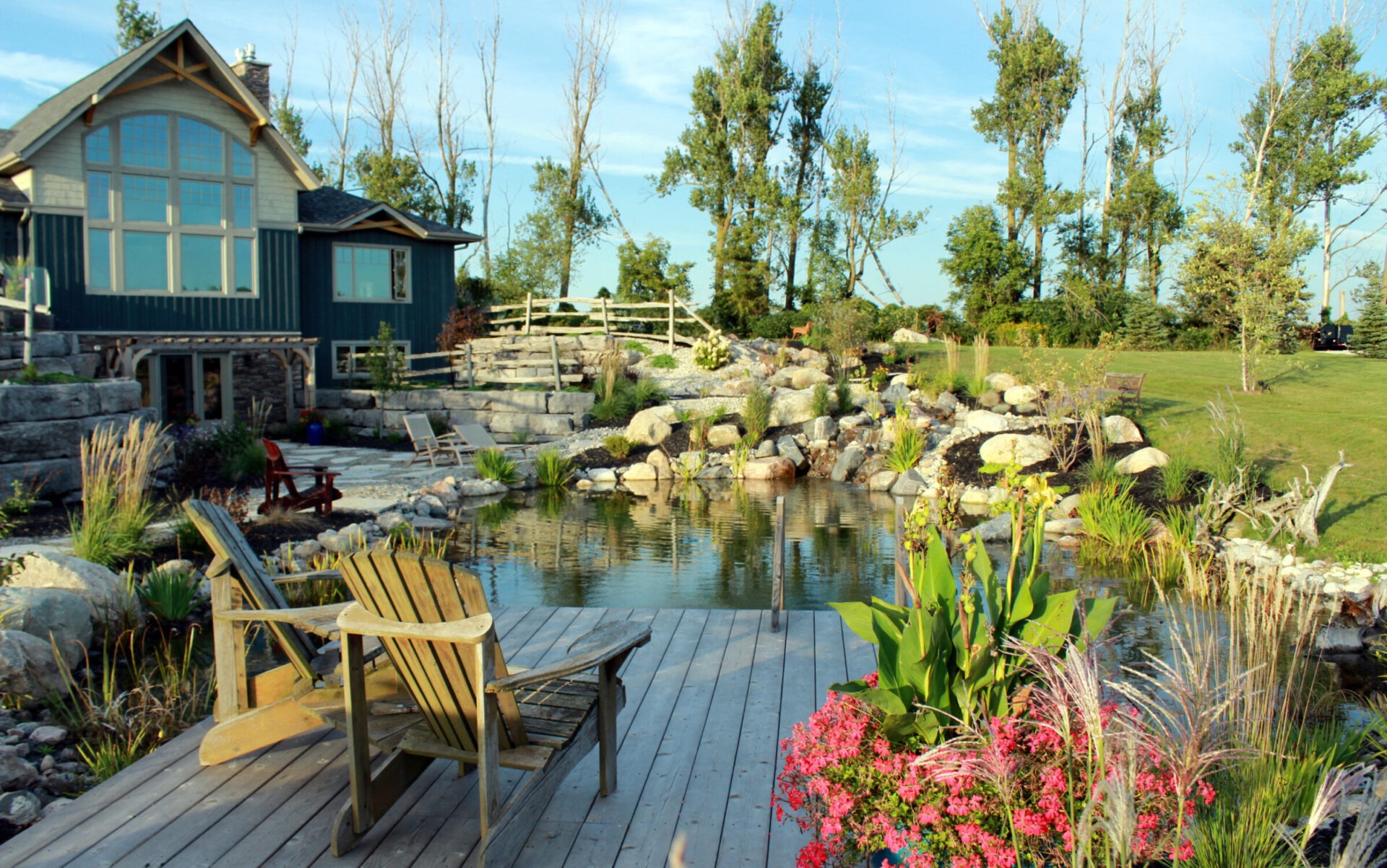 A tranquil backyard with a pond, wooden chairs on a deck, colorful flowers, a stone pathway, landscaped with rocks, trees, and a blue house.