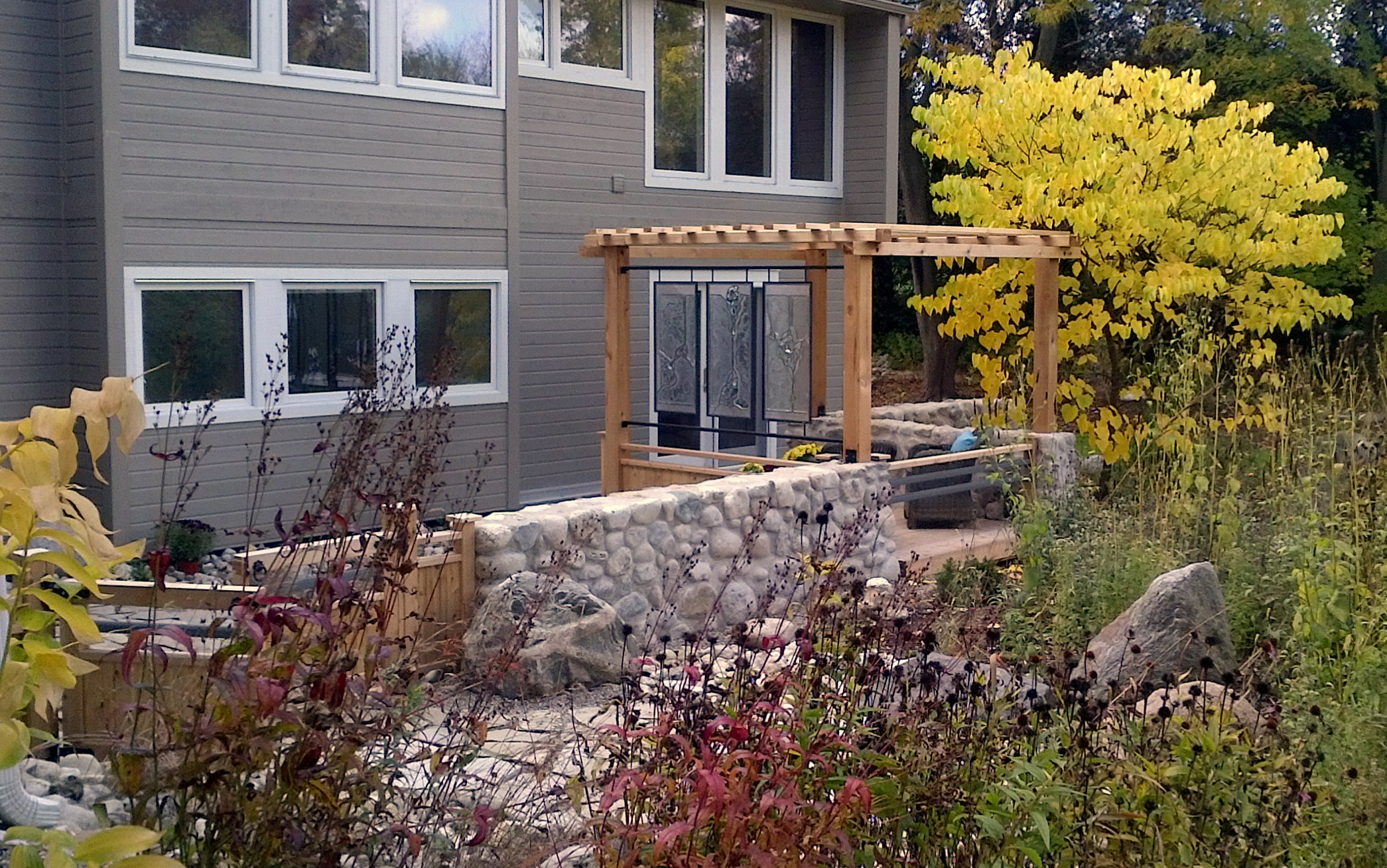 A modern house with large windows, surrounded by a landscaped garden with a wooden pergola, stone retaining walls, and colorful autumn foliage.