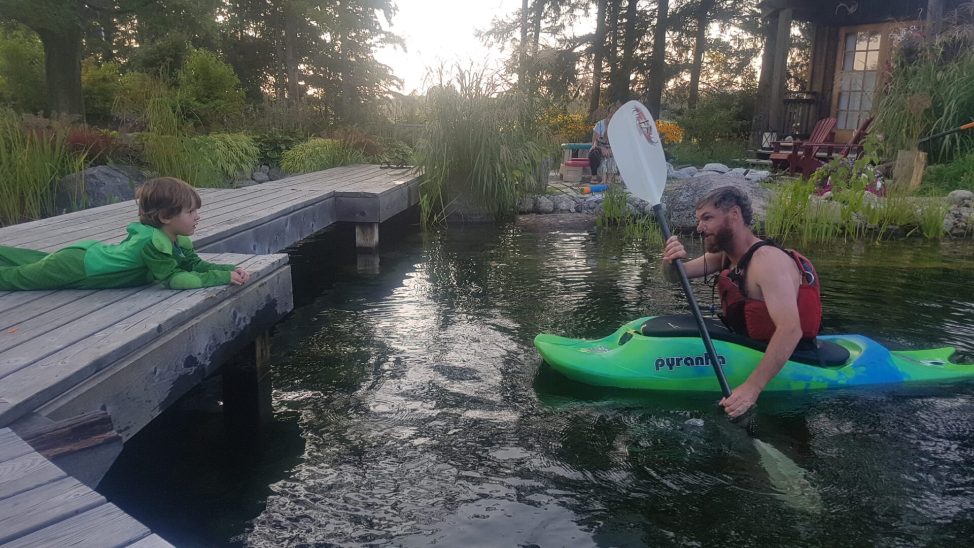 A child lies on a wooden dock beside a calm pond, watching a person in a green kayak paddling near lush garden and a cozy house.