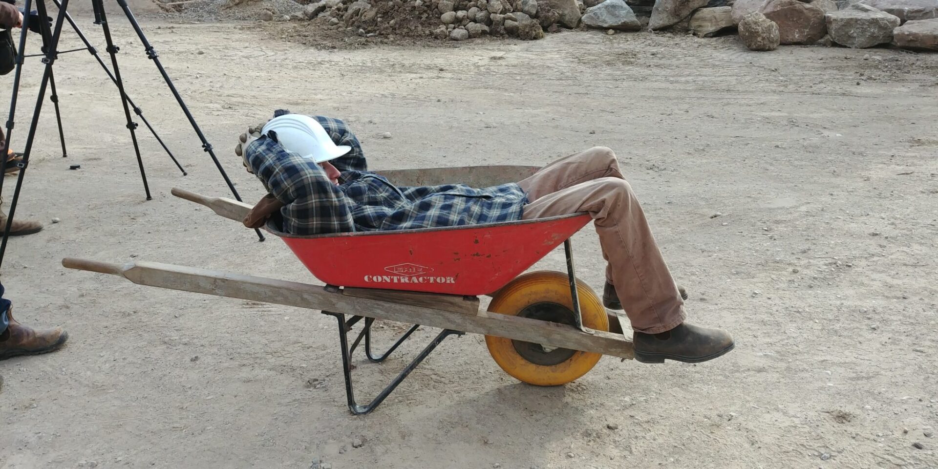 A person is reclining in a red wheelbarrow, wearing a plaid shirt, brown pants, a white hard hat, and work boots, at a construction site.