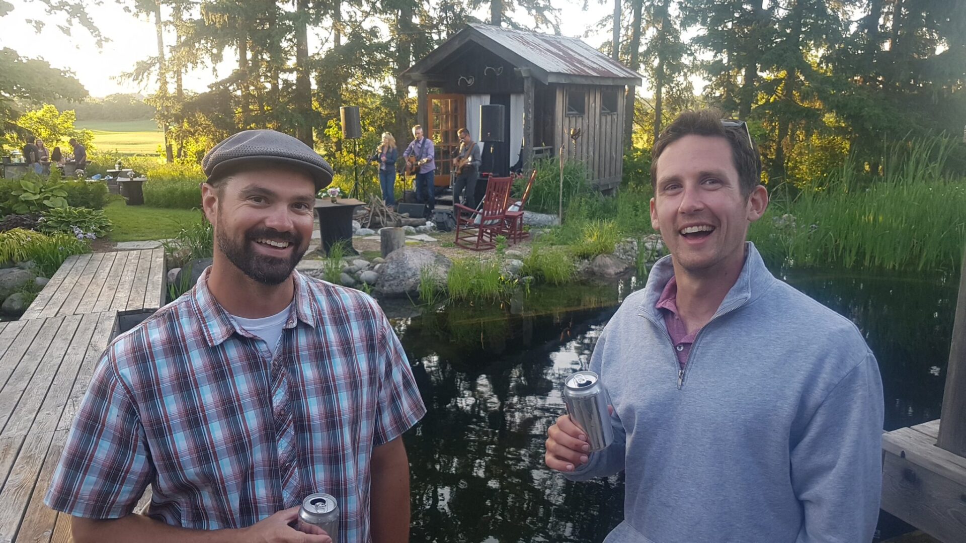 Two people holding drinks smile by a pond with a small hut and a band playing in the background during an outdoor gathering in daylight.