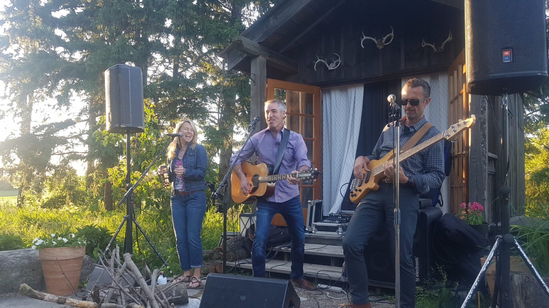 Three people are performing outdoors on a small stage, surrounded by trees at sunset. They have a guitar, bass guitar, and microphone setup.