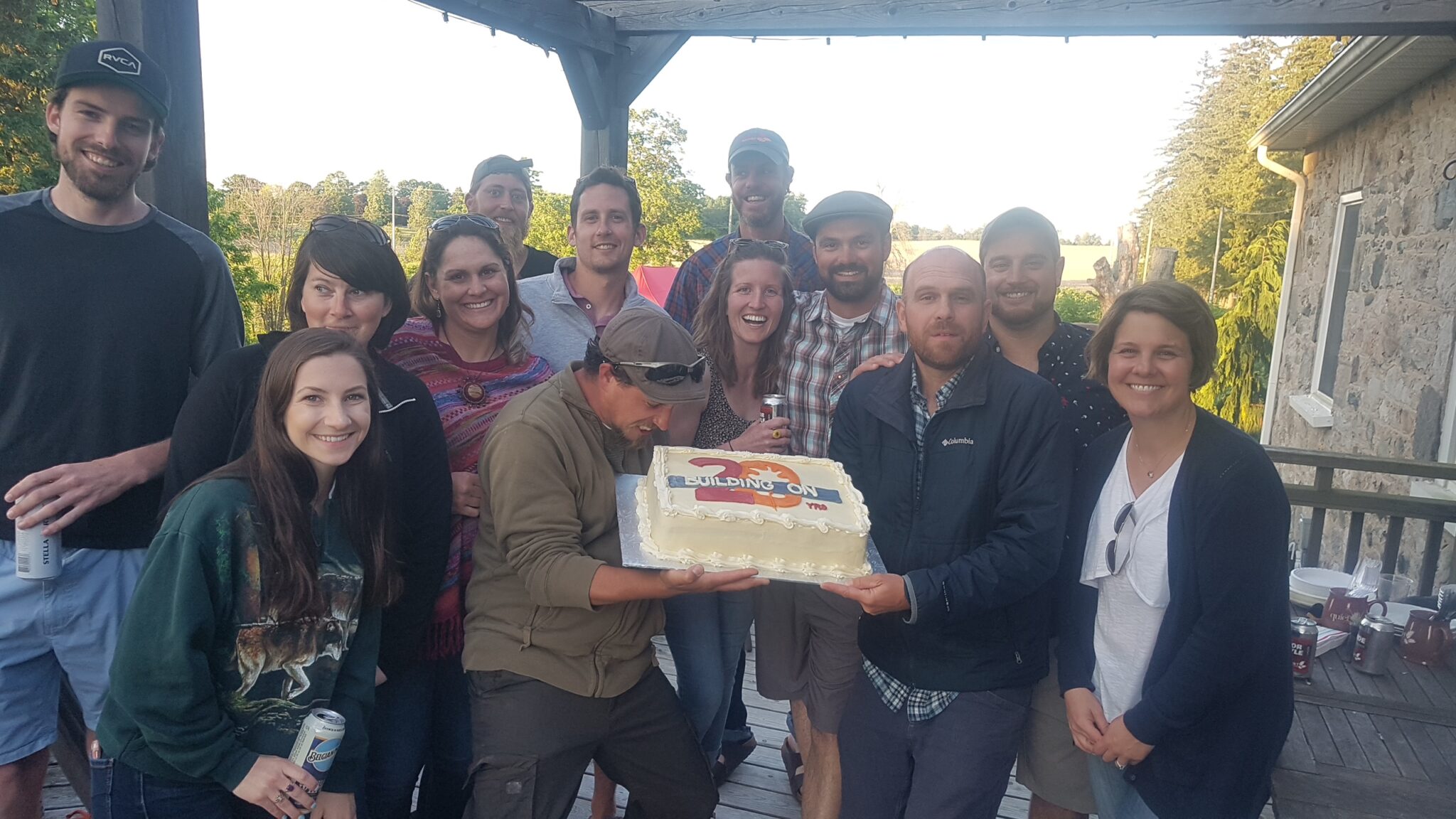 A group of people posing on a porch with smiles, one person in front holding a large cake that says "Building on 70" for a celebration.