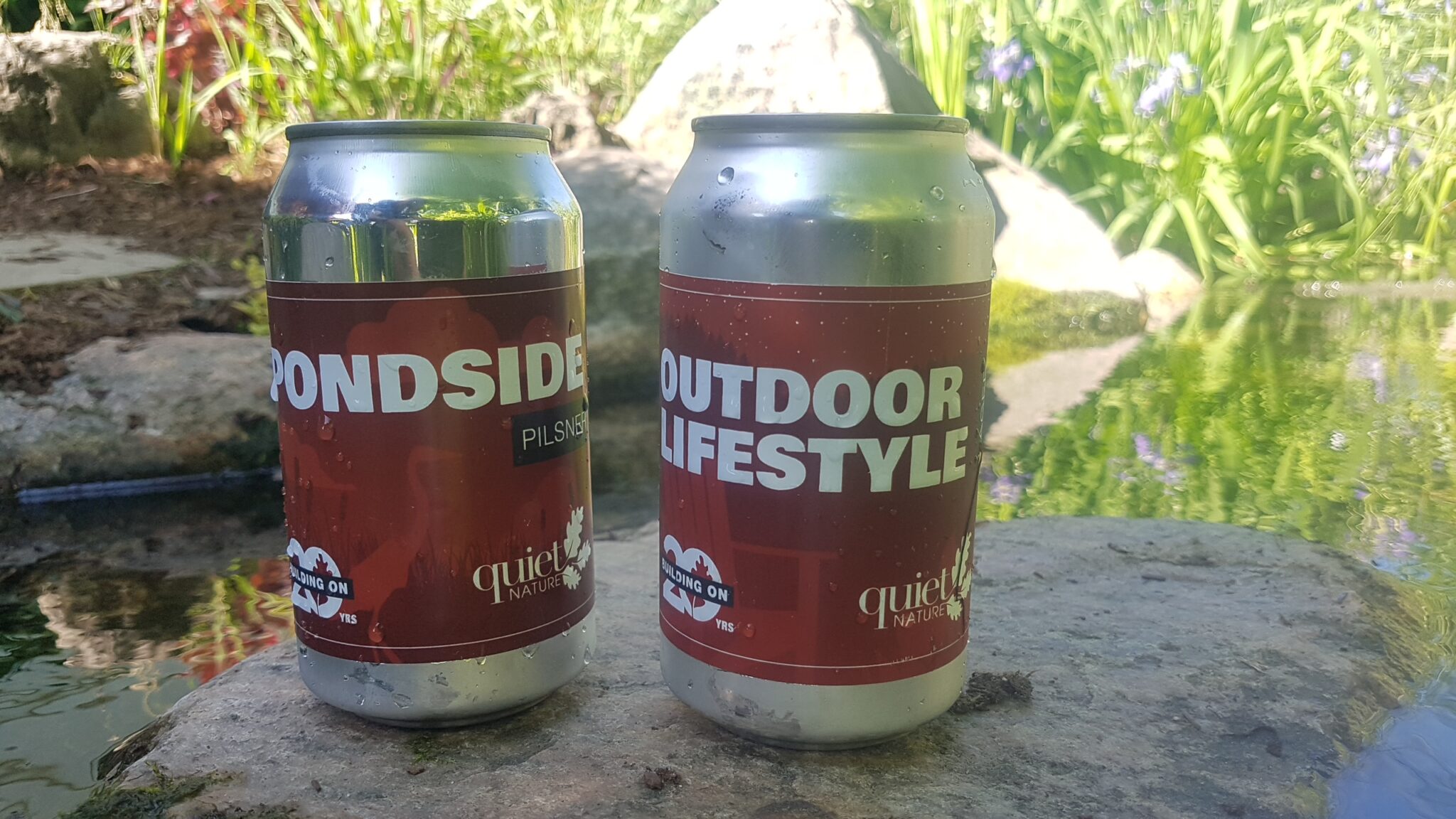 Two cans labeled "PONDSIDE" and "OUTDOOR LIFESTYLE" rest on a rock beside a pond, surrounded by greenery and reflections on the water.