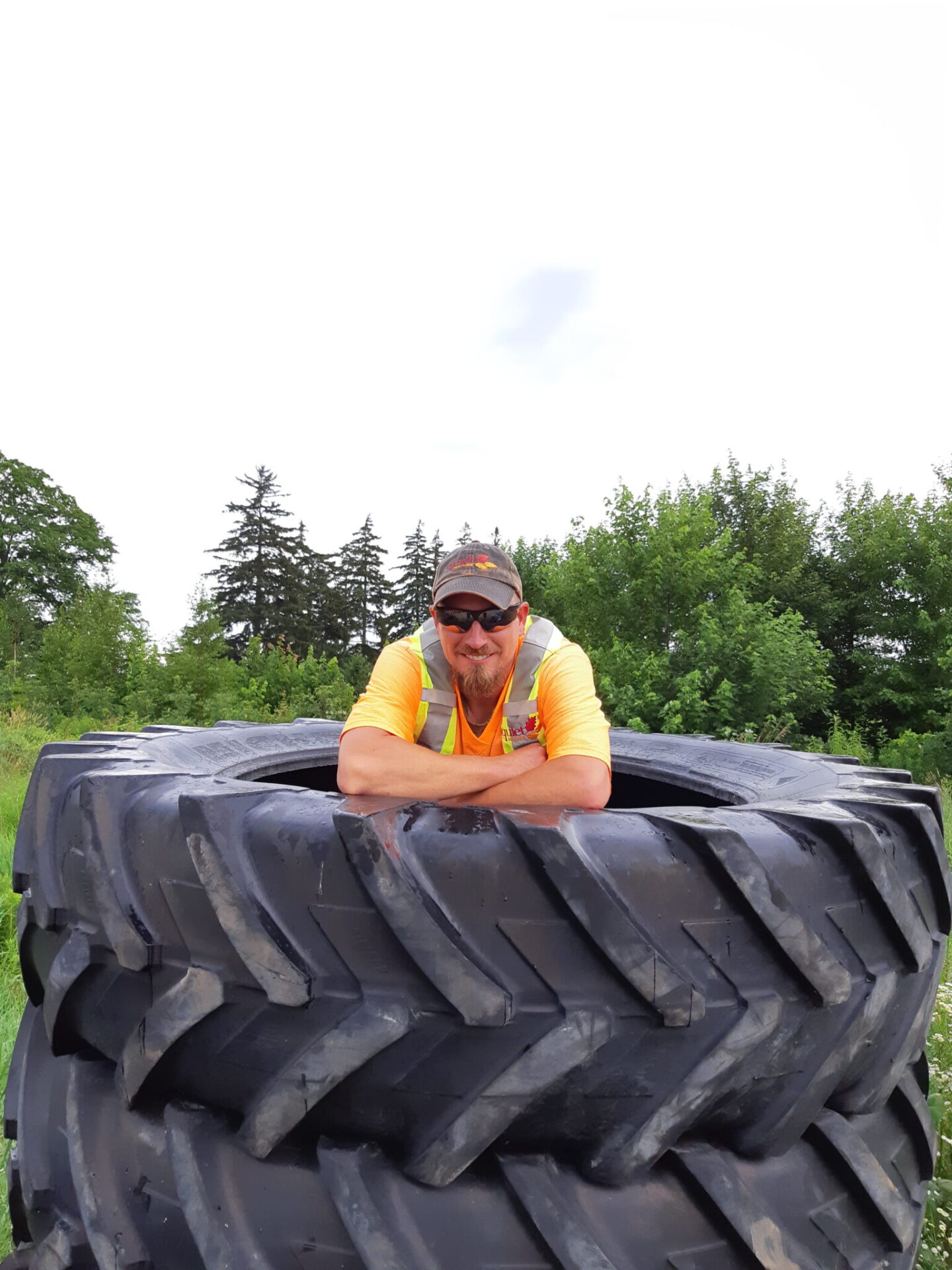 A smiling person in a high-visibility shirt leans on a large tractor tire outdoors, with trees and a cloudy sky in the background.