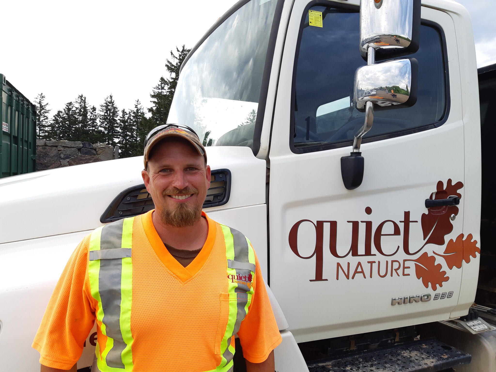 A smiling person wearing a high-visibility vest stands in front of a white truck with "Quiet Nature" and leaf graphics on it, outdoors under a clear sky.