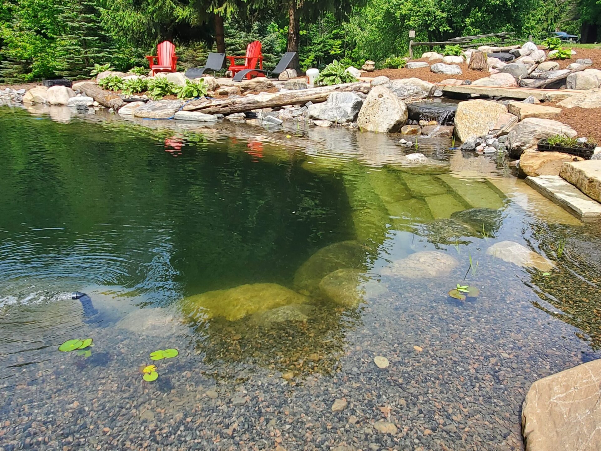 This image shows a tranquil pond surrounded by rocks, with red chairs on the bank, lush greenery, and a small waterfall in a landscaped garden.