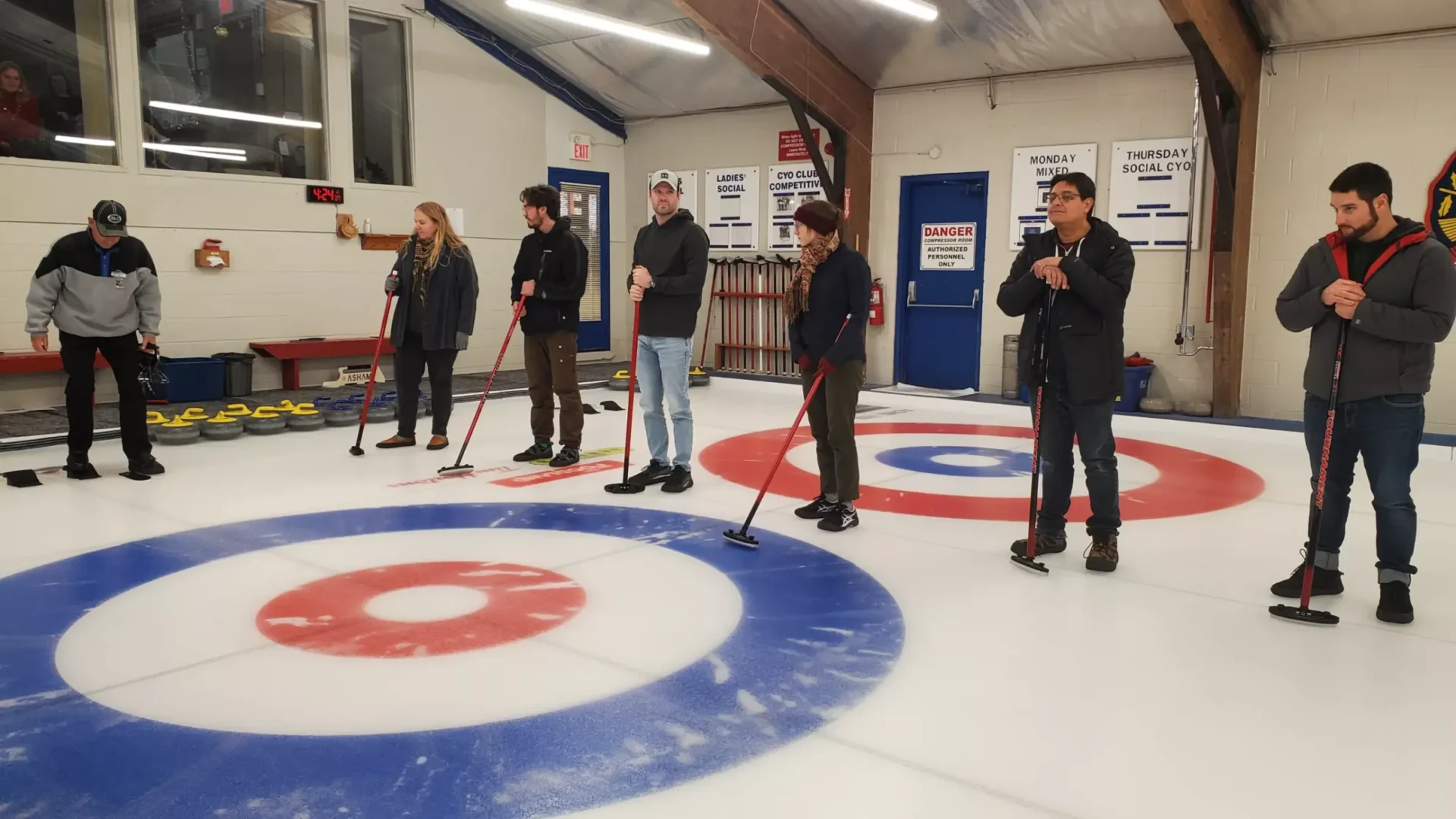 People stand on a curling rink, holding brooms, with curling stones in the background. They appear to be learning the game or preparing to play.