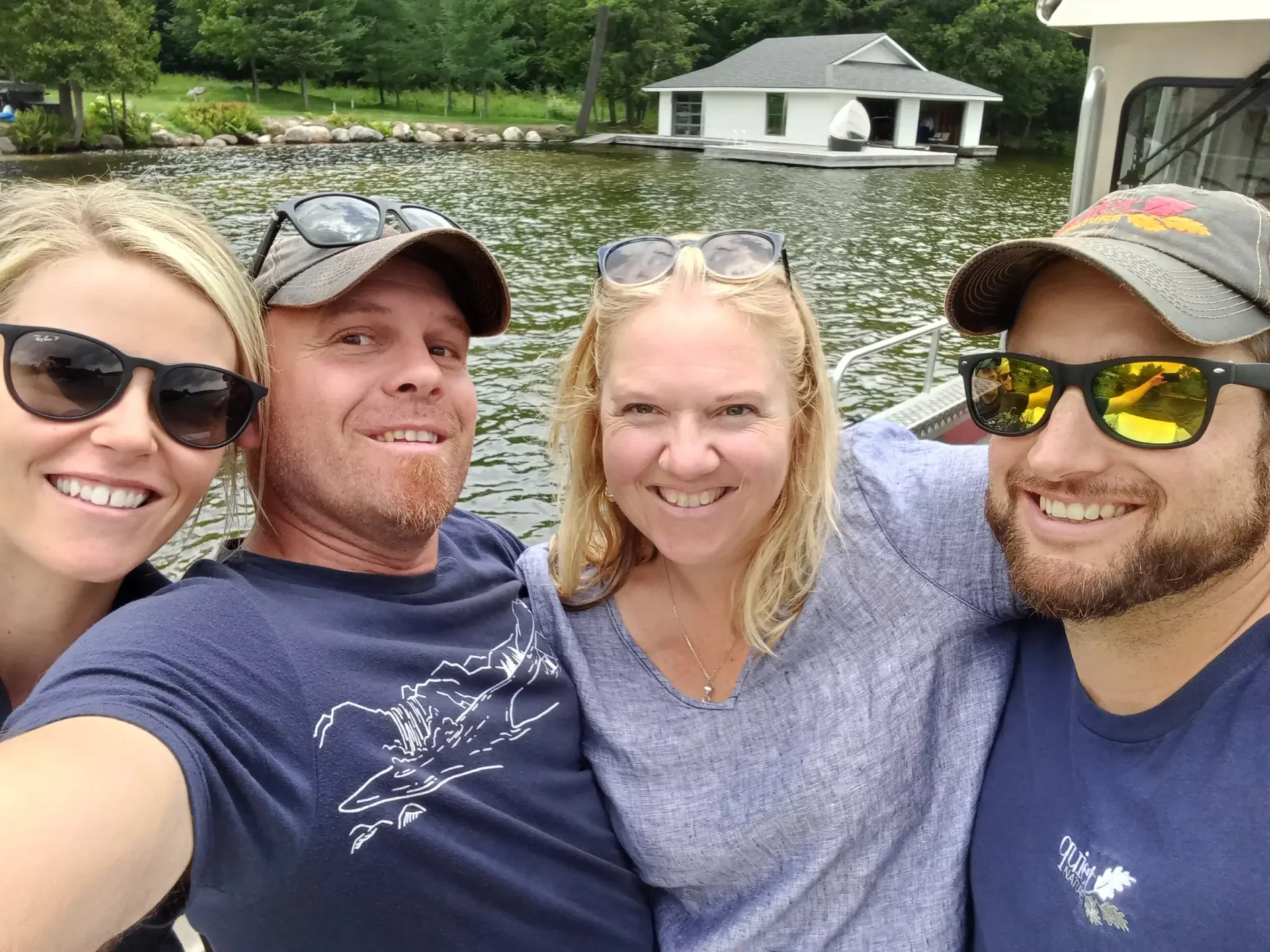 Four smiling people are taking a selfie on a boat, with sunglasses on, a lake and a boathouse in the background. They appear to be enjoying their time together.