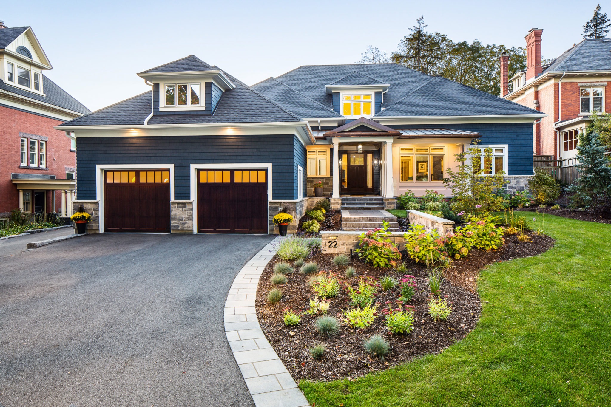 A luxurious house with a stone pathway, a double garage, well-manicured garden, at dusk with interior and exterior lights on.