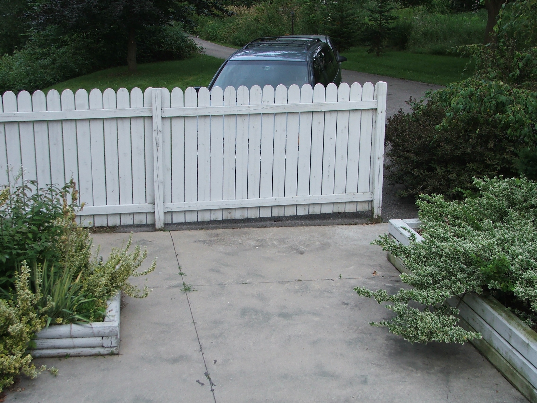 A white picket fence with a swing gate is shown. In the background, there's a car parked beside lush greenery. Concrete driveway and planter boxes visible.