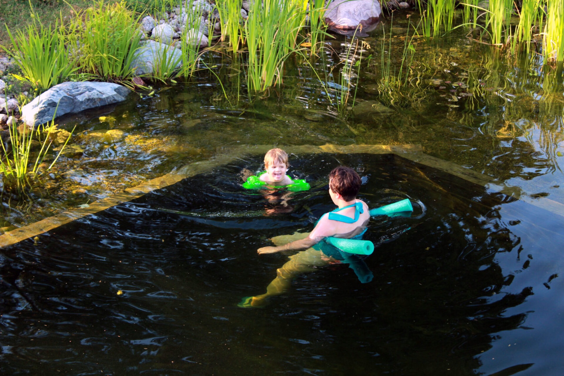 A person and a child are enjoying time in a natural pond surrounded by green plants and stones, both using floatation devices for safety.