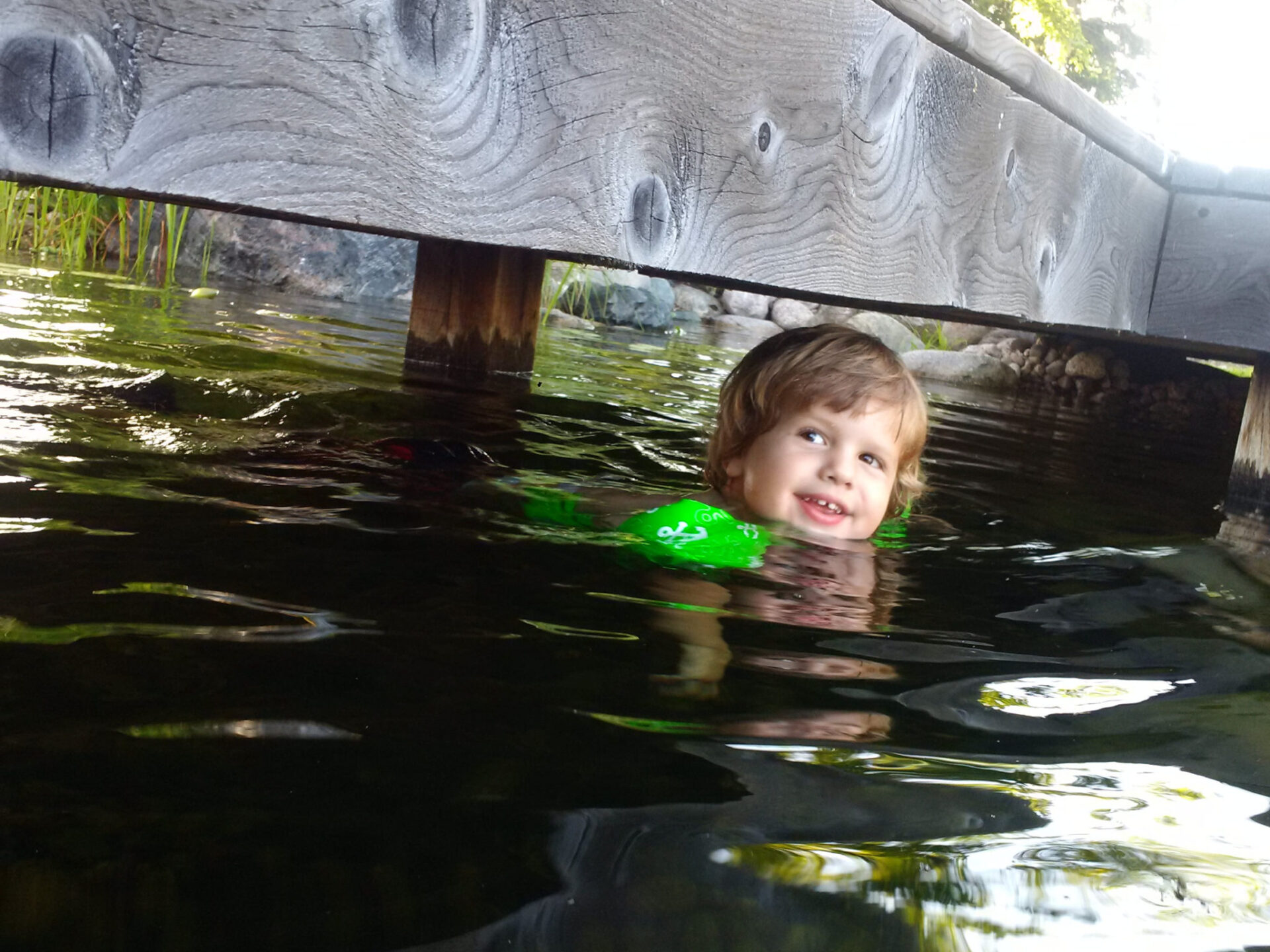 A young child with a joyful expression swims under a wooden structure in a body of water, wearing a green floatation device with a frog design.