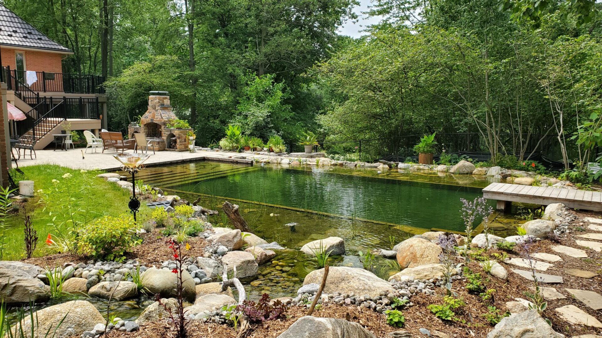 A serene backyard with a natural-looking pond surrounded by rocks, plants, a path, an outdoor fireplace, seating area, and dense trees in the background.