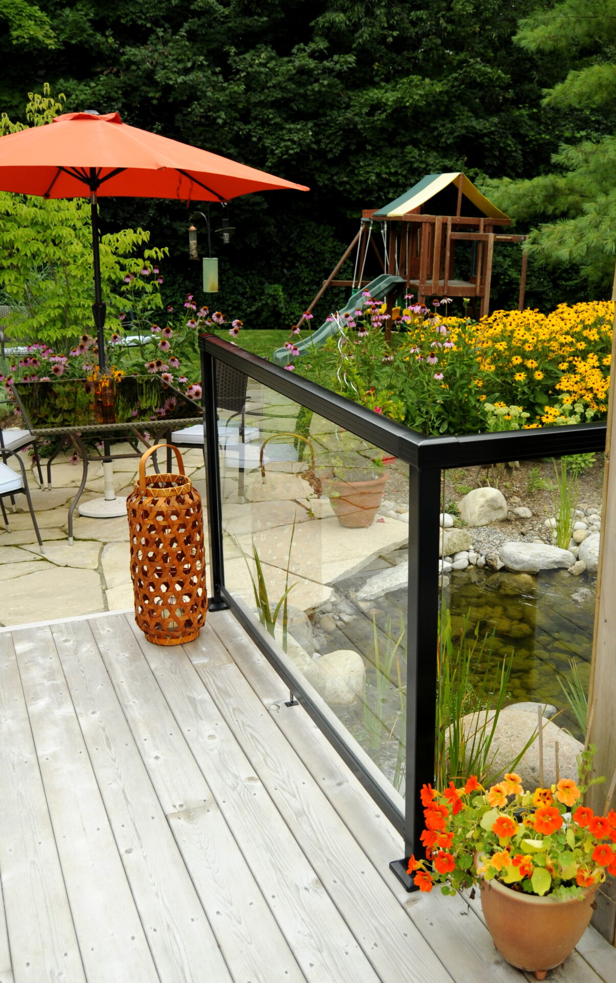 A serene outdoor deck with a glass railing overlooks a garden with colorful flowers, a playground, and a pond. A woven lantern accentuates the space.