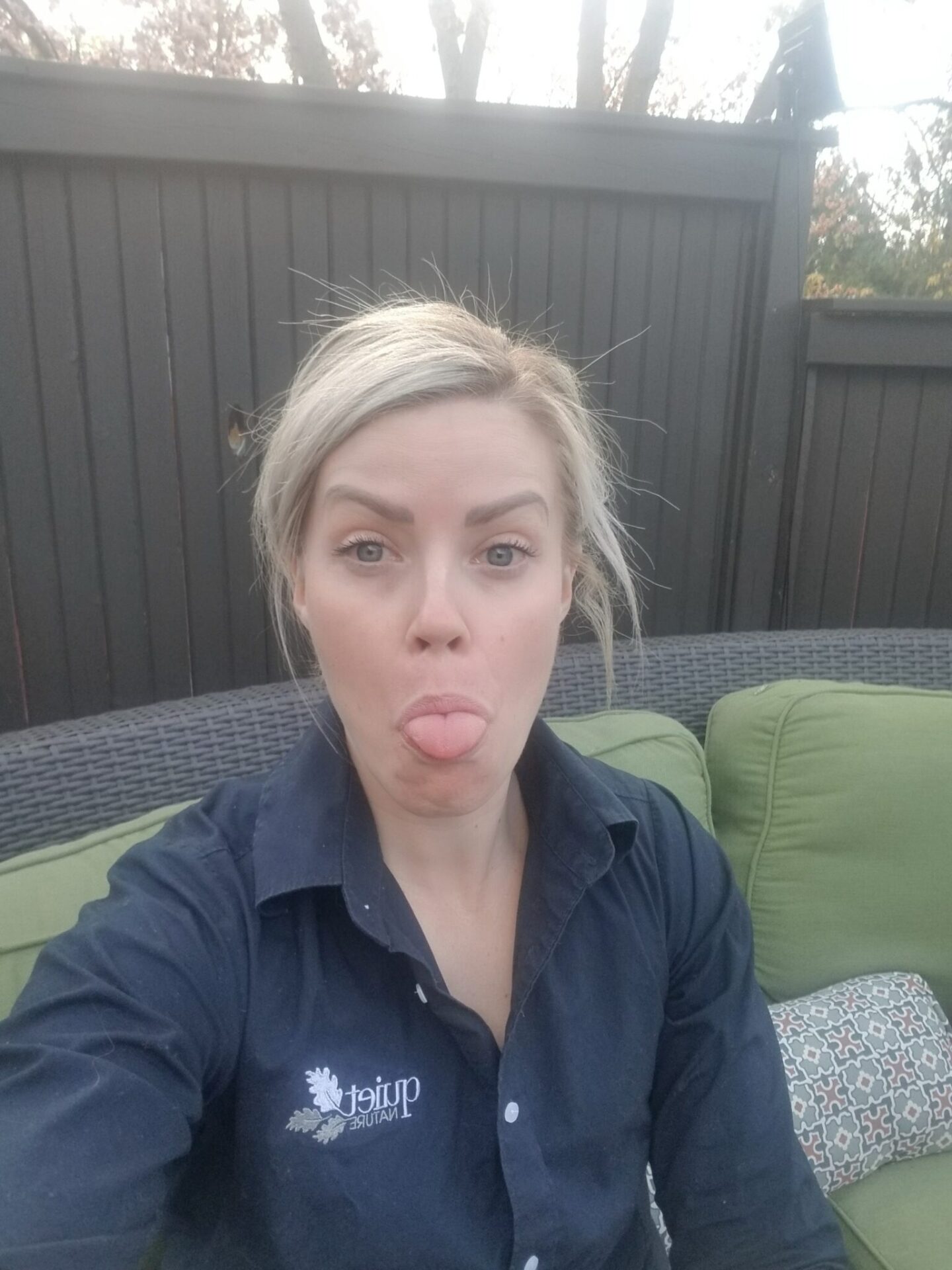 A person is making a playful face with their tongue out, sitting on a green patio couch in front of a dark fence, wearing a dark work shirt.