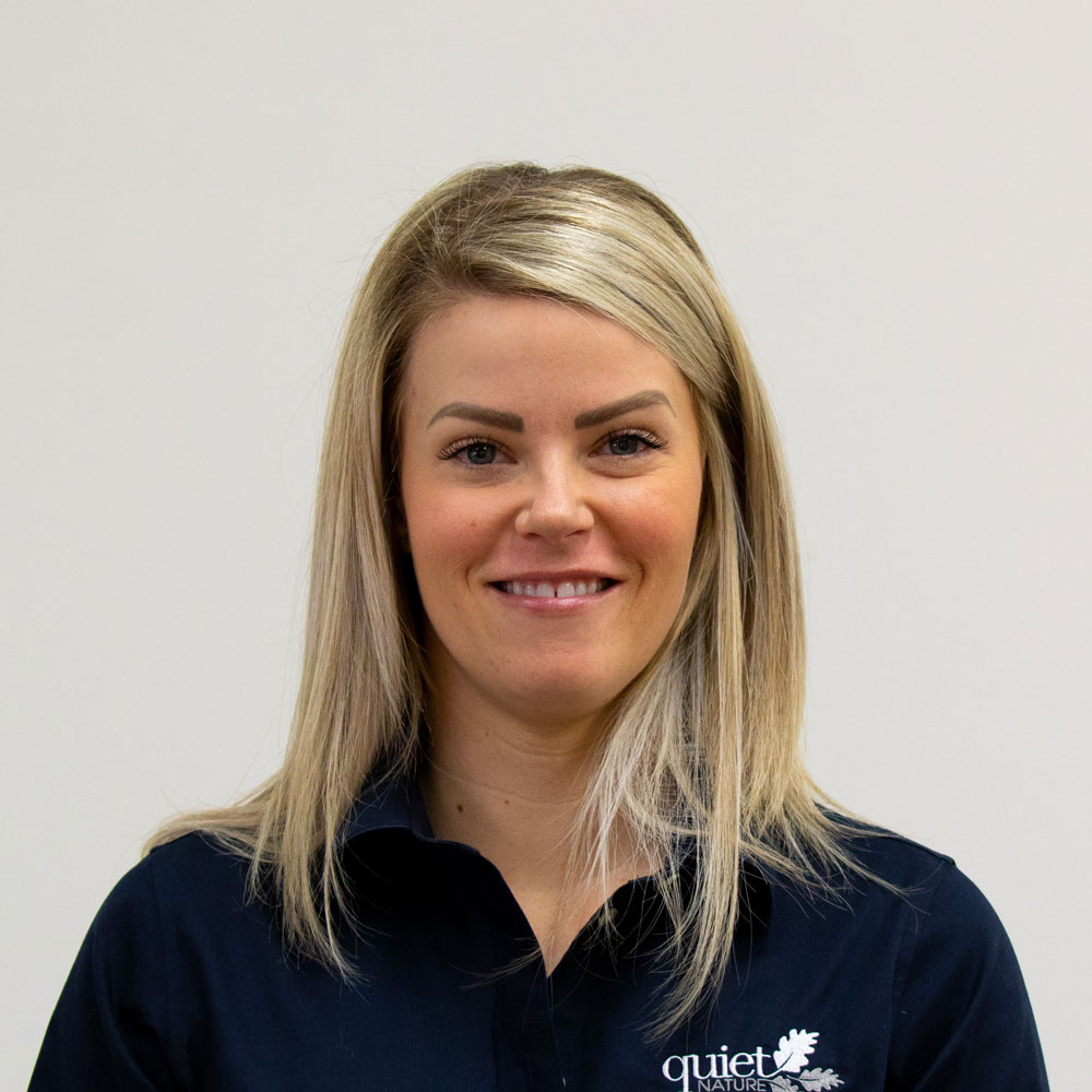 A person is smiling at the camera, wearing a dark blue shirt with the logo "quiet nature." They have short blond hair and a light background.